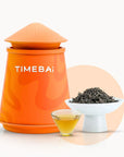 Timebai da hong pao oolong tea with an orange tea canister, loose leaves in a white bowl, and a glass cup of golden tea on a white background.