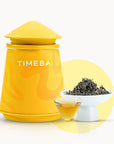 Timebai gardenia dancong oolong tea with a yellow tea canister, loose leaves in a white bowl, and a glass cup of golden tea on a white background.