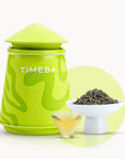 Timebai jasmine mao jian green tea with a lime green tea canister, loose leaves in a white bowl, and a glass cup of pale yellow tea on a white background.